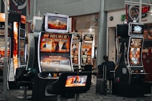 A collection of slot machines is situated inside a building with bright, colorful screens displaying various games and gaming themes. A person is seated nearby, seemingly relaxed with luggage beside them, and the atmosphere is lively with electronic displays.