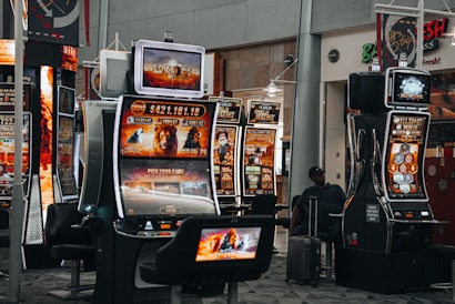 A collection of slot machines is situated inside a building with bright, colorful screens displaying various games and gaming themes. A person is seated nearby, seemingly relaxed with luggage beside them, and the atmosphere is lively with electronic displays.