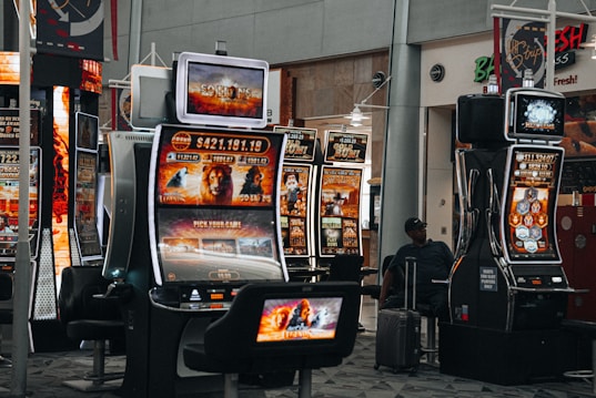 A collection of slot machines is situated inside a building with bright, colorful screens displaying various games and gaming themes. A person is seated nearby, seemingly relaxed with luggage beside them, and the atmosphere is lively with electronic displays.