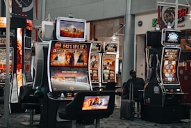 A collection of slot machines is situated inside a building with bright, colorful screens displaying various games and gaming themes. A person is seated nearby, seemingly relaxed with luggage beside them, and the atmosphere is lively with electronic displays.