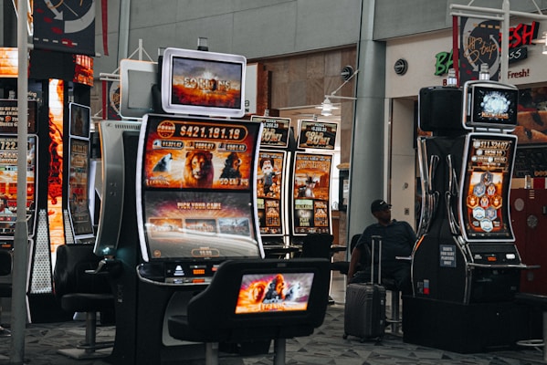 A collection of slot machines is situated inside a building with bright, colorful screens displaying various games and gaming themes. A person is seated nearby, seemingly relaxed with luggage beside them, and the atmosphere is lively with electronic displays.