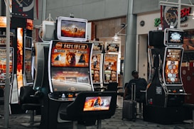 A collection of slot machines is situated inside a building with bright, colorful screens displaying various games and gaming themes. A person is seated nearby, seemingly relaxed with luggage beside them, and the atmosphere is lively with electronic displays.