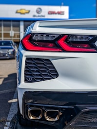 A close-up view of the rear end of a modern sports car, showcasing its sleek taillights and dual exhaust system. The car is parked in a dealership lot, seen with signage for Chevrolet, Buick, and GMC in the background.