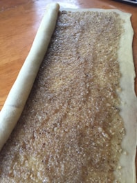 A sheet of dough partially rolled up, with a textured brown filling spread evenly across its surface, likely indicating it is being prepared for baking.