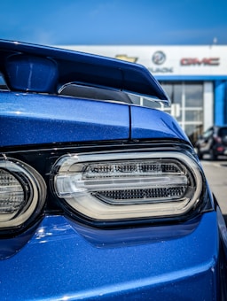 A close-up view of the rear of a blue car, focusing on its taillight and spoiler. The background shows an automotive dealership with logos for Chevrolet, Buick, and GMC, indicating a car sales setting.