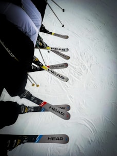 A group of skiers is lined up with their skis on a snowy surface. The skis are of the brand 'HEAD' and vary in colors, including blue, red, and yellow accents. Ski poles are visible, indicating readiness for skiing.