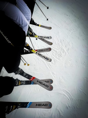 A group of skiers is lined up with their skis on a snowy surface. The skis are of the brand 'HEAD' and vary in colors, including blue, red, and yellow accents. Ski poles are visible, indicating readiness for skiing.