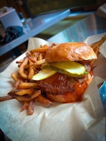 A juicy burger with pickles and a glossy bun is served with a side of golden, crispy fries. The burger appears to have a drizzle of sauce, adding to its appetizing look. The background consists of a metallic table and chairs, suggesting a casual dining setting.