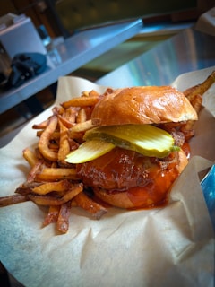 A juicy burger with pickles and a glossy bun is served with a side of golden, crispy fries. The burger appears to have a drizzle of sauce, adding to its appetizing look. The background consists of a metallic table and chairs, suggesting a casual dining setting.