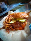 A juicy burger with pickles and a glossy bun is served with a side of golden, crispy fries. The burger appears to have a drizzle of sauce, adding to its appetizing look. The background consists of a metallic table and chairs, suggesting a casual dining setting.