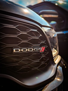 A close-up view of a car's front grille with a prominent 'DODGE' emblem. The grille features a hexagonal pattern, and the metallic surface reflects the surrounding light.