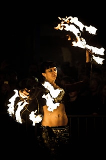 A performer is twirling fire poi, creating striking patterns of light against a dark background. Her outfit is adorned with intricate designs, and she appears focused and skillful in her movements.