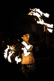 A performer is twirling fire poi, creating striking patterns of light against a dark background. Her outfit is adorned with intricate designs, and she appears focused and skillful in her movements.