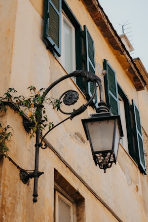 An old-fashioned metal street lamp mounted on the side of a weathered yellow wall. The building has green shutters and an overgrown vine creeping around the lamp. The architecture reflects a rustic, historical aesthetic.
