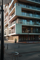 A modern, multi-story apartment building with large glass balconies and reflective windows. The structure features clean lines and an industrial aesthetic. A person wearing a green jacket and dark pants walks along the sidewalk next to the building. The scene is bathed in soft, late afternoon light, casting long shadows on the ground.