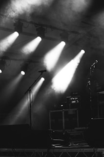 A dimly lit stage environment features several spotlights beaming down through a haze of smoke, creating a dramatic atmosphere. Microphones stand ready, and music equipment is visible in the background, suggesting a setup for a live performance.