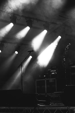 A dimly lit stage environment features several spotlights beaming down through a haze of smoke, creating a dramatic atmosphere. Microphones stand ready, and music equipment is visible in the background, suggesting a setup for a live performance.