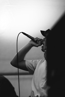 A person wearing a hat and a t-shirt is passionately singing into a microphone. The scene is captured in black and white, emphasizing the intensity of the performance. The microphone cable is visible, and the setting appears to be indoors.