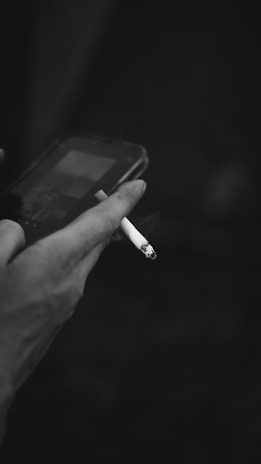 A hand is holding a cigarette while interacting with a smartphone. The focus is on the hand and the lit cigarette, with smoke visibly emitting. The image is in black and white, providing a moody and somewhat contemplative atmosphere.
