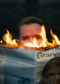 A person is seen looking over a newspaper that is partially engulfed in flames. The fire creates a dramatic contrast against the neutral background and highlights, emphasizing intensity and focus in the subject's eyes.