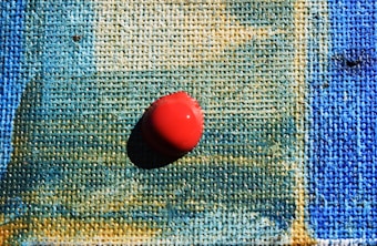 A close-up of a textured canvas with shades of blue and green blending together, featuring a single red pushpin placed prominently in the center, casting a shadow.