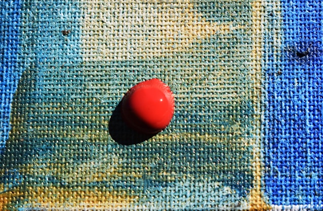 A close-up of a textured canvas with shades of blue and green blending together, featuring a single red pushpin placed prominently in the center, casting a shadow.