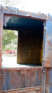 A rusty, old industrial container with visible weathering, showing an open side revealing the inside. Yellow cylindrical objects are stacked inside. In the background, trees are visible, and a red bench with a patterned cloth is seen on a concrete surface.