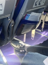 An airplane seat with safety instructions visible on the back. Below the seat, a pair of black shoes and gray socks are resting on the purple carpeted floor. A mesh pocket holds some reading material.