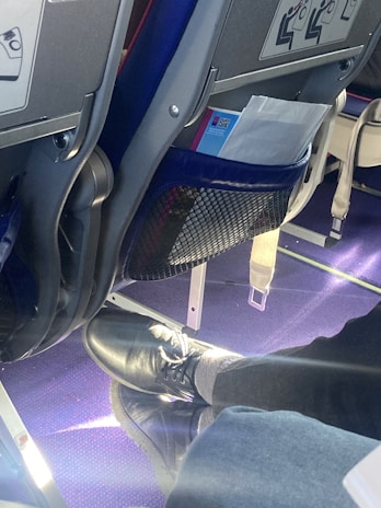 An airplane seat with safety instructions visible on the back. Below the seat, a pair of black shoes and gray socks are resting on the purple carpeted floor. A mesh pocket holds some reading material.