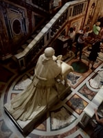 A marble statue of a robed figure kneels at a prayer bench in an ornate setting. The floor and walls are adorned with intricate geometric and floral patterns. Several people are observing or taking pictures, some in motion, within a dimly lit, opulent interior.