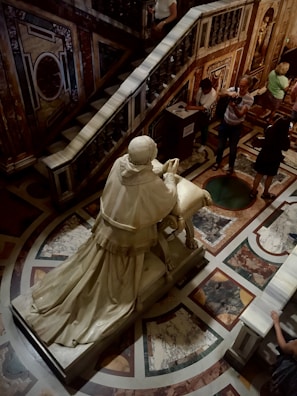 A marble statue of a robed figure kneels at a prayer bench in an ornate setting. The floor and walls are adorned with intricate geometric and floral patterns. Several people are observing or taking pictures, some in motion, within a dimly lit, opulent interior.