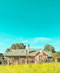 A charming countryside house featuring a green roof and brick walls, surrounded by a garden full of vibrant flowers and greenery. The sky is clear and blue with a few scattered clouds, adding to the serene and picturesque setting. Trees frame the house, enhancing the natural ambiance.