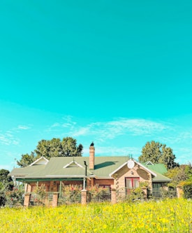 A charming countryside house featuring a green roof and brick walls, surrounded by a garden full of vibrant flowers and greenery. The sky is clear and blue with a few scattered clouds, adding to the serene and picturesque setting. Trees frame the house, enhancing the natural ambiance.