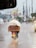 A motorcycle is carrying a large load covered with a tarp along a wet road. The background features an urban setting with vehicles, trees, utility poles, and a billboard visible through the rain.