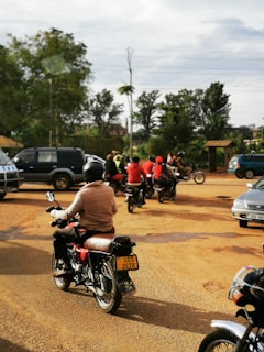 Several motorcyclists are riding on a dirt road, surrounded by a few cars. The scene is set outdoors with trees in the background. The riders are wearing helmets and are in casual clothing. The road is wide, and the sky is cloudy.