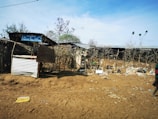 A makeshift structure made of corrugated metal and wood stands in front of a dry, barren landscape. Sparse vegetation and scattered debris are visible on the ground. A sign hanging from the structure reads 'Welcome to Kudero Fuel Station.'