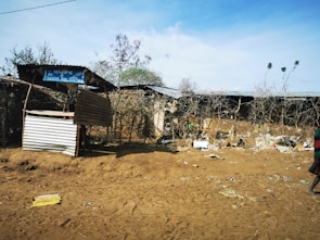 A makeshift structure made of corrugated metal and wood stands in front of a dry, barren landscape. Sparse vegetation and scattered debris are visible on the ground. A sign hanging from the structure reads 'Welcome to Kudero Fuel Station.'