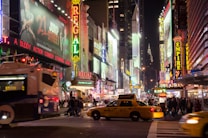 A vibrant city scene featuring bustling streets filled with people, illuminated by bright neon signs and billboards. Yellow taxis and a city bus highlight the urban environment. The atmosphere is lively, with advertisements for movies and entertainment venues creating a dynamic and colorful backdrop.