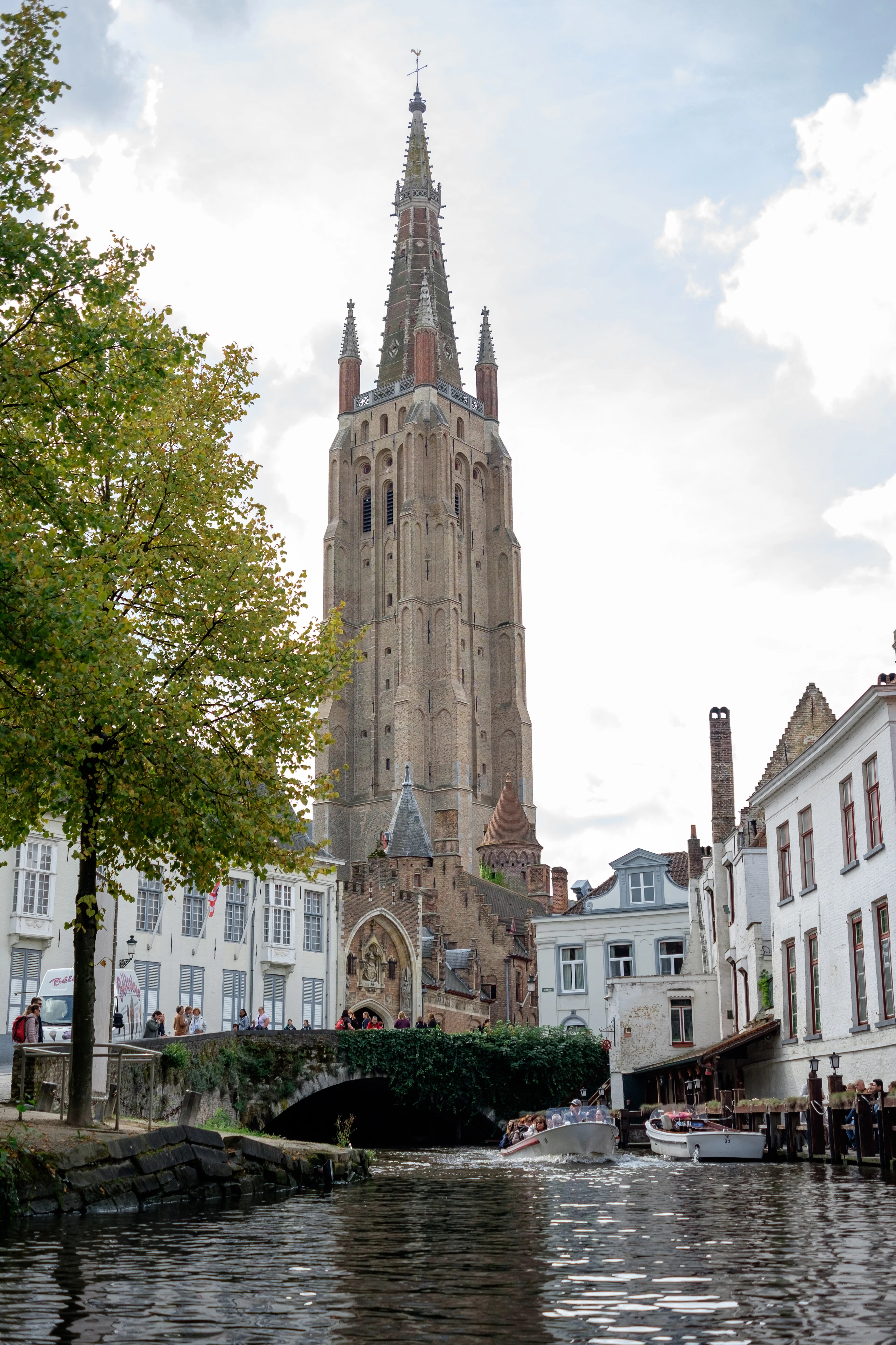 The brick spire of the Church of Our Lady towers over a canal bridge as tour boats pass below.