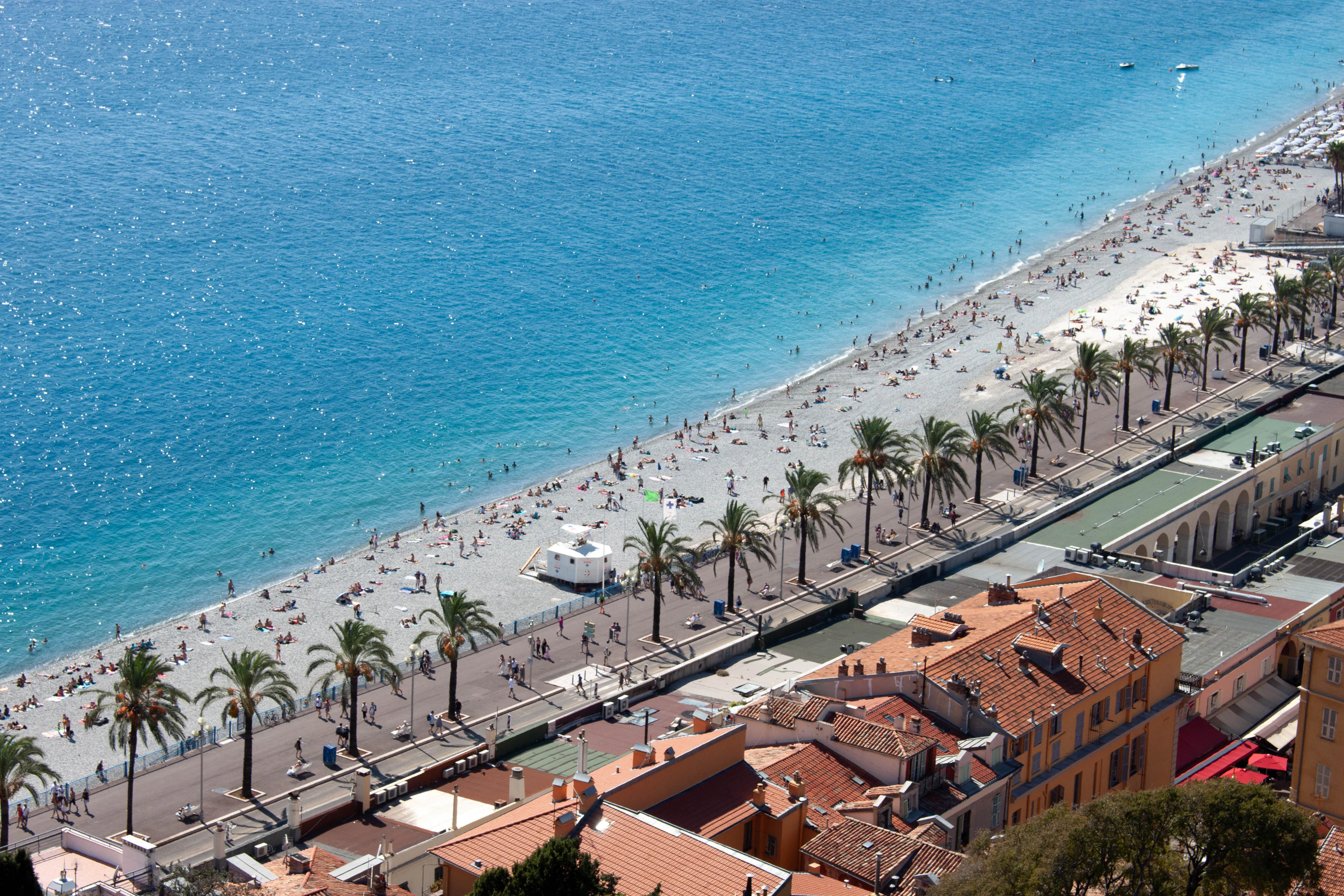 Looking down on the palm-lined Promenade des Anglais in Nice and the pebble beach of the Baie des Anges, with swimmers in the turquoise water.
