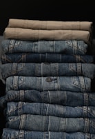 A neatly stacked collection of folded pants, with two khaki colored pants at the top followed by multiple layers of blue denim jeans below.