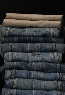 A neatly stacked collection of folded pants, with two khaki colored pants at the top followed by multiple layers of blue denim jeans below.