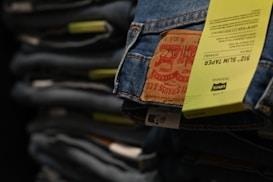 A close-up view of a stack of jeans, focusing on one pair with a prominent leather patch and a yellow tag indicating the size and fit details. The jeans appear to be neatly folded on a shelf, suggesting a retail or storage setting.