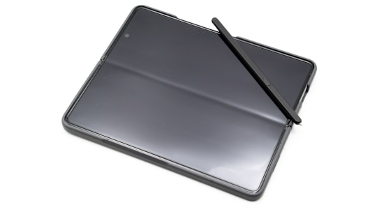 A sleek black tablet with a foldable screen, accompanied by a black stylus placed diagonally across it. The surface of the tablet is glossy, reflecting light, and it features minimalist design elements.