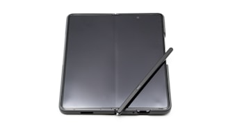 A foldable smartphone lies open on a flat surface with a stylus resting diagonally across its screen. The device features a seamless, continuous display with a slim profile, indicating modern design and advanced technology.