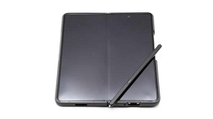 A foldable smartphone lies open on a flat surface with a stylus resting diagonally across its screen. The device features a seamless, continuous display with a slim profile, indicating modern design and advanced technology.