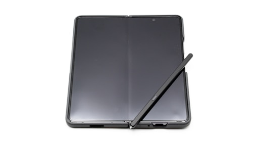 A foldable smartphone lies open on a flat surface with a stylus resting diagonally across its screen. The device features a seamless, continuous display with a slim profile, indicating modern design and advanced technology.