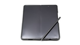 A foldable smartphone lies open on a flat surface with a stylus resting diagonally across its screen. The device features a seamless, continuous display with a slim profile, indicating modern design and advanced technology.