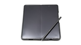 A foldable smartphone lies open on a flat surface with a stylus resting diagonally across its screen. The device features a seamless, continuous display with a slim profile, indicating modern design and advanced technology.