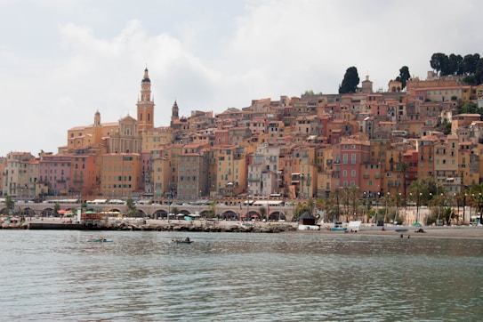 A picturesque coastal town with a cluster of colorful, historic buildings overlooking the water. The architecture features warm hues of yellow, pink, and orange, creating a charming and inviting appearance. Tall trees and a church tower rise above the buildings, adding to the scenic view. The calm waterfront is dotted with people and small boats.